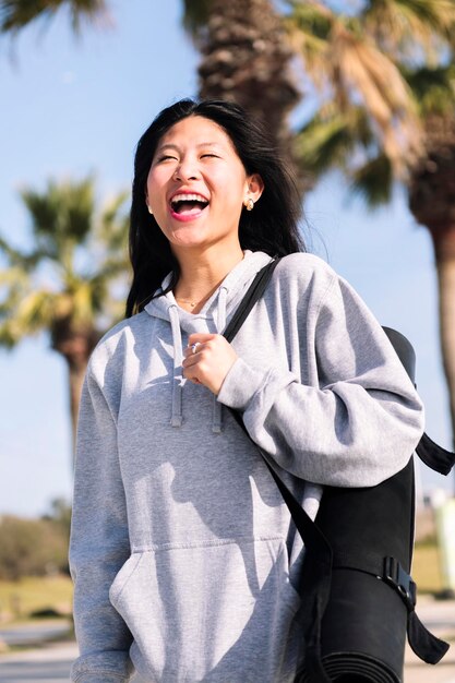 Eine asiatische Frau lacht, während sie mit einer Yoga-Matte spazieren geht
