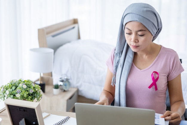 Foto eine asiatische frauenerkrankung brustkrebspatientin mit rosa schleife mit kopftuch nach der behandlung zur chemotherapie mit arbeitsgeschäft am laptop im büro zu hause, medizinkonzept