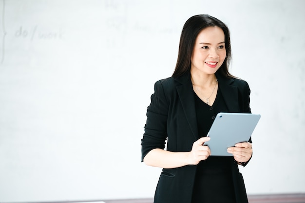 Eine asiatische Lehrerin mittleren Alters, die ein Tablet im Whiteboard des Klassenzimmers an der Universität hält