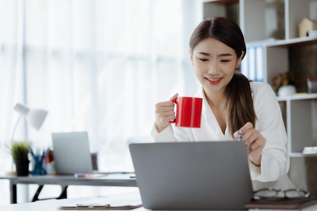 Eine asiatische Mitarbeiterin sitzt mit einer roten Kaffeetasse im Büro der Marketingabteilung des Startups. Sie ist Marketing-Kundenkontakt- und Beratungsmitarbeiterin Marketingkonzept