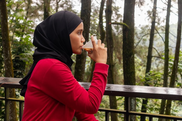 Eine asiatische muslimische Frau genießt eine Plastiktasse Eiskaffee auf dem Balkon mit Naturbäumen im Hintergrund