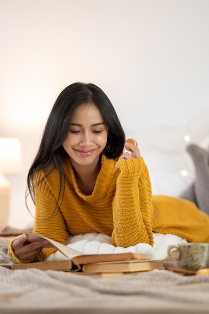 Eine attraktive asiatische Frau in einem kuscheligen Pullover isst gerne Snacks und liest ein Buch auf ihrem Bett