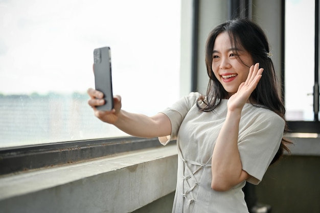 Eine attraktive asiatische Frau macht Selfies mit ihrem Smartphone, während sie am Fenster steht