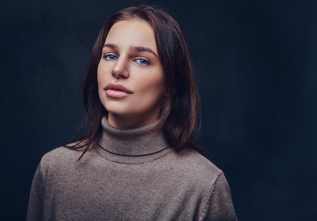 Eine attraktive brünette Frau in einer braunen Jacke mit langem Hals über grauem Hintergrund.