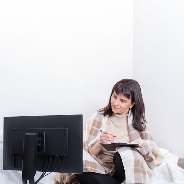 Eine attraktive Frau bedeckt mit einer Decke, die ein Notizbuch und einen Stift hält und einen Computermonitor zu Hause genau betrachtet