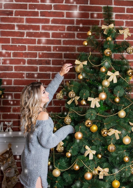 Eine attraktive Frau in schickem Strickkleid schmückt im Winter einen Weihnachtsbaum für die Feier zu Hause