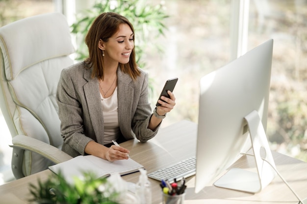 Eine attraktive Geschäftsfrau, die Smartphone verwendet, während sie in ihrem Heimbüro am Computer arbeitet.