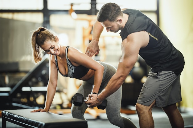 Eine attraktive junge Frau, die mit einer Hantel unter der Kontrolle des persönlichen Trainers im Fitnessstudio trainiert.