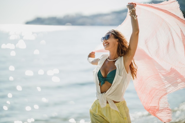 Eine attraktive junge Frau hat Spaß und genießt einen Sommerurlaub. Sie posiert mit transparentem Schal in ihren Händen am Strand.