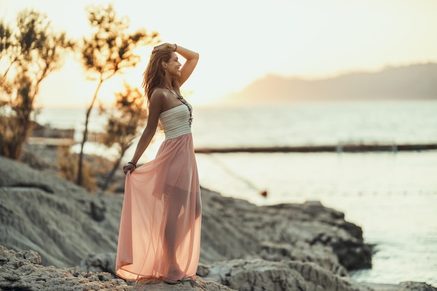 Eine attraktive junge Frau im Kleid genießt den Sommersonnenuntergang an der wunderschönen Küste.