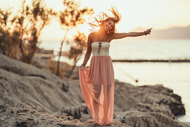 Eine attraktive junge Frau im Sommerkleid hat Spaß, während sie den Sonnenuntergang an der wunderschönen Küste verbringt.