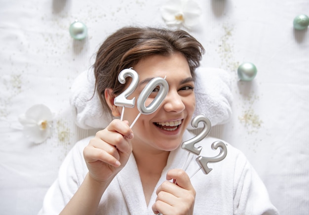 Eine attraktive junge Frau liegt in einem weißen Gewand für eine Kurbehandlung, hält die Zahlen 2022 auf weißem Hintergrund, das Konzept von Weihnachten und Neujahr.