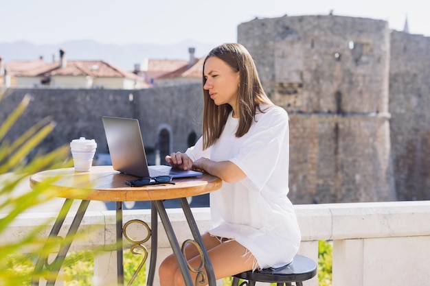 Eine attraktive junge Frau sitzt an einem Tisch vor dem Café und arbeitet an einem Laptop