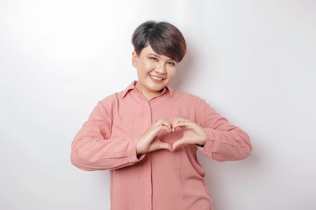 Eine attraktive junge kurzhaarige asiatische Frau, die ein rosa Hemd trägt, fühlt sich glücklich und eine romantische Herzgeste drückt zarte Gefühle aus