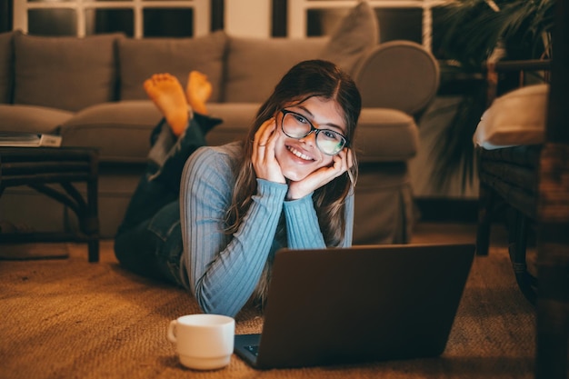 Eine attraktive kaukasische und junge Frau mit Brille mit Computer auf dem Teppich zu Hause in der Nacht Schöne Dame entspannt Surfen im Netz mit Blick auf die cameraxA