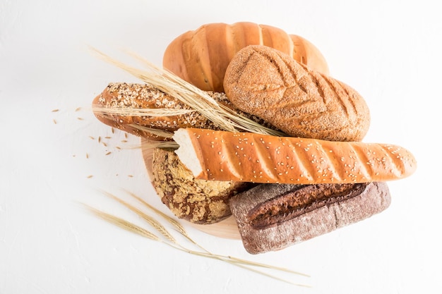 Eine Auswahl an gebackenem Brot aus verschiedenen Mehlen auf einem Holzbrett und einem weißen Hintergrund mit Ähren aus Weizen und Flachs und Sesamsamen