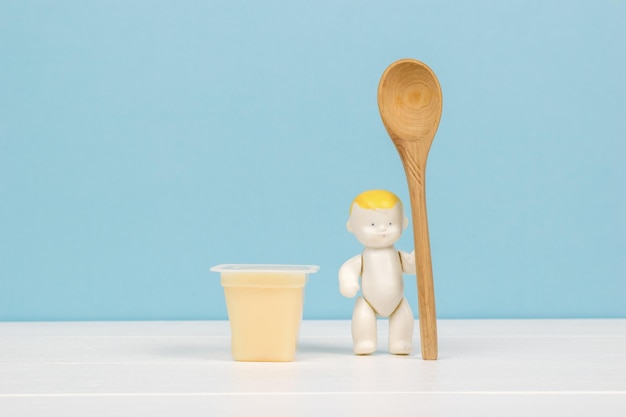 Eine Babyfigur mit einem Holzlöffel und einer Tasse Bananendessert auf einem Holztisch. Bananen-Dessert. Das Konzept der gesunden Babynahrung.