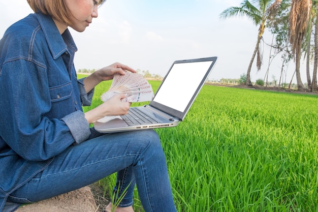 Eine Bäuerin, die in einem Feld mit Banknoten sitzt und einen Laptop verwendet.