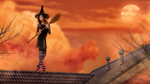 Eine Ballerina in Spitzenschuhen in einem schwarzen Hexenkostüm mit Hut und Besen tanzt auf dem Dach eines alten Hauses vor dem Hintergrund eines bedrohlich hellen Sonnenuntergangs