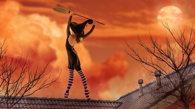 Eine Ballerina in Spitzenschuhen in einem schwarzen Hexenkostüm mit Hut und Besen tanzt auf dem Dach eines alten Hauses vor dem Hintergrund eines bedrohlich hellen Sonnenuntergangs