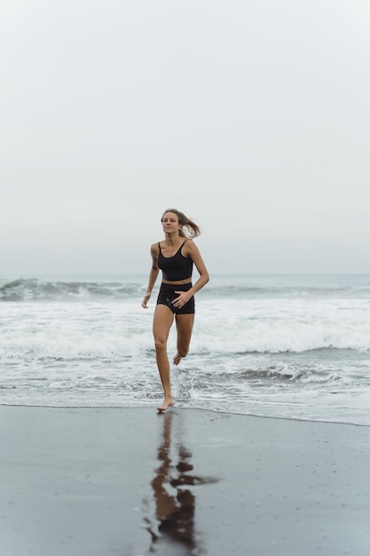 Eine barfüßige junge Frau mit einem schlanken Körper läuft auf der Meeresbrandung am Wasserbecken, um sich fit zu halten und Fett zu verbrennen. Strandhintergrund mit blauem Himmel. Frauenfitness, Joggen, sportliche Aktivitäten auf einer Sommerfami