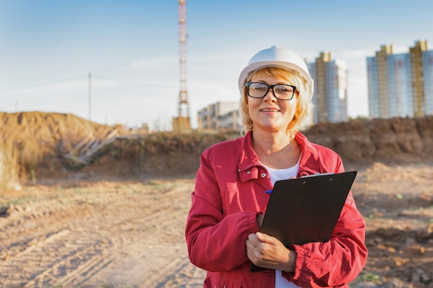 Eine Bauarbeiterin ist auf einer Baustelle. Eine Frau mittleren Alters in einem weißen Helm mit einem Tablet macht eine Inspektion des Territoriums.