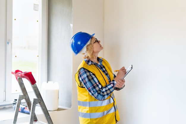 Eine Baumeisterin mit einem Tablet in der Hand inspiziert einen Raum in einem Neubau. Frau mittleren Alters mit Helm macht Reparaturen in der Wohnung.