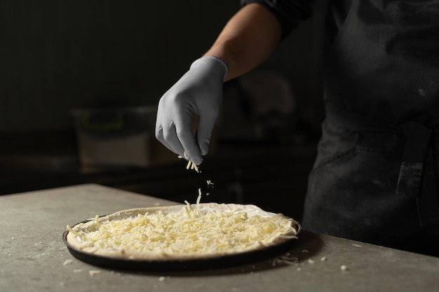 Eine behandschuhte Hand streut geriebenen harten Mozzarella-Käse auf eine runde rohe Pizza