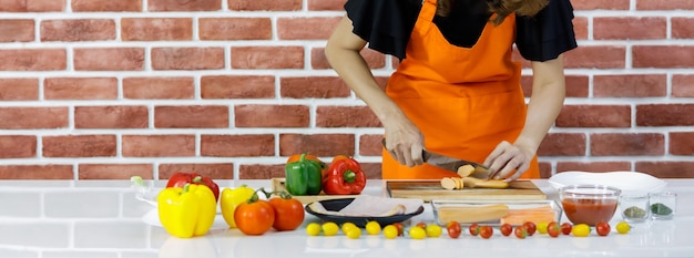 Eine beschnittene Frau in orangefarbener Schürze steht vor einer braunen Backsteinwand der Küche und schneidet sorgfältig Hotdog auf einem Kochtisch, der voller frischer Tomaten, gesunder Paprika und Geschirr ist