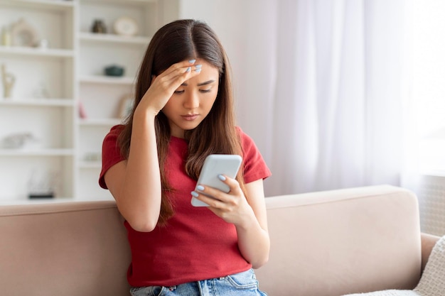 Eine besorgte junge asiatische Frau berührt ihre Stirn, während sie eine Nachricht auf ihrem Handy liest