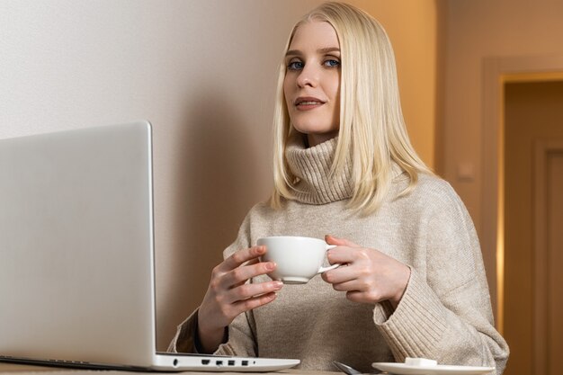 Eine blauäugige Blondine in einem beigen Pullover mit weitem Kragen sitzt mit einer weißen Tasse Tee am Computer