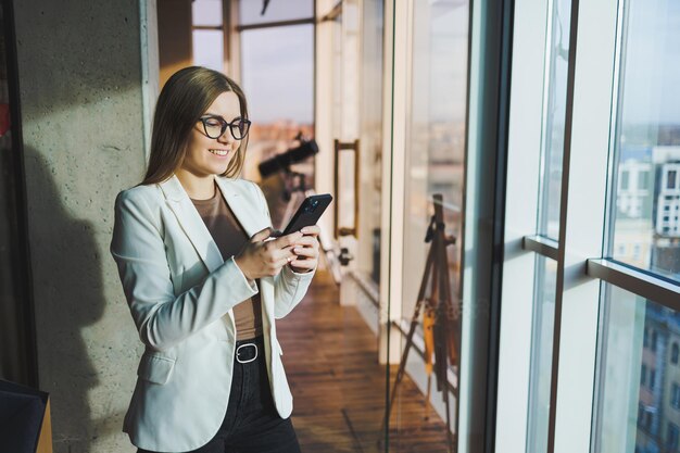 Eine blonde Europäerin in weißer Jacke und Brille telefoniert, während sie in einem geräumigen Büro mit großen Fenstern steht Erfolgreiche junge Freiberuflerin