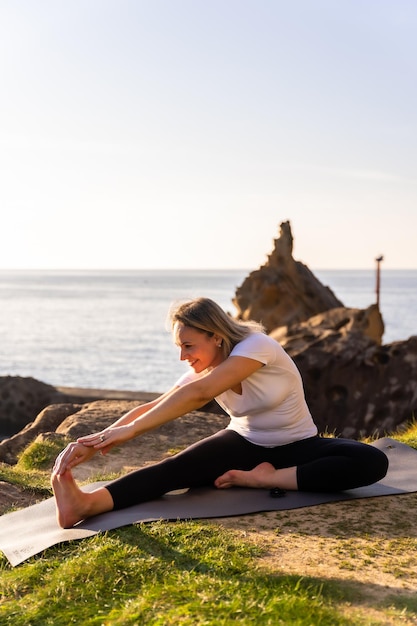 Eine blonde Frau, die Yoga-Übungen in der Natur am Meer macht, das sich neben einem Leuchtturm erstreckt