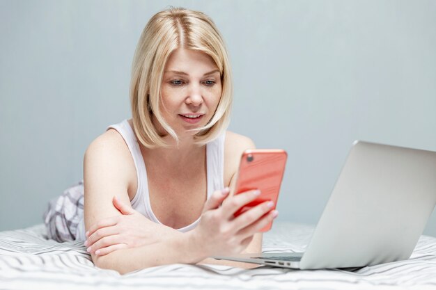 Eine blonde Frau in Hauskleidung liegt mit einem Smartphone in der Hand und einem Laptop auf einem Bett. Fernarbeit während der Coronavirus-Pandemie.