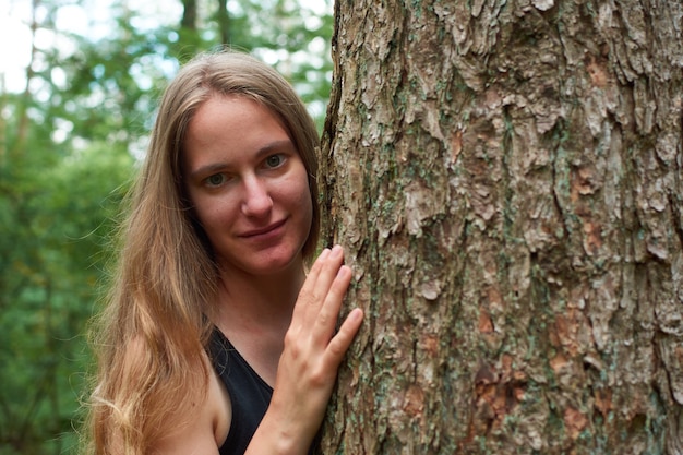 Eine blonde kaukasische Dame mit langen Haaren, die hinter einem Baumstamm hervorschaut und lächelt