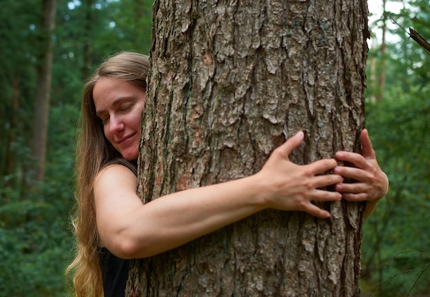 Eine blonde kaukasische Dame mit langen Haaren umarmt einen Baum im Wald