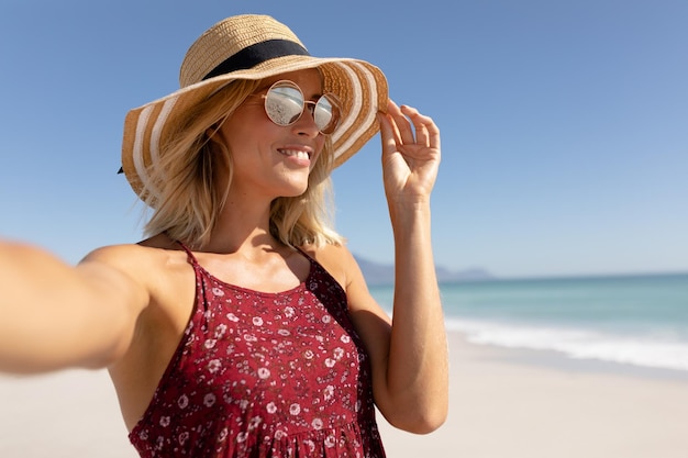 Eine blonde kaukasische Frau mit Strohhut und Sonnenbrille genießt im Urlaub ihre Freizeit an einem sonnigen Strand, hält ihren Hut in der Hand, lächelt und macht ein Selfie mit blauem Himmel und Meer im Hintergrund