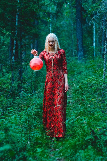 Eine Blondine in einem roten Kleid im Wald mit einer roten chinesischen Laterne in ihren Händen