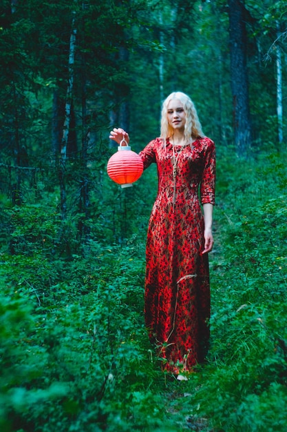 Eine Blondine in einem roten Kleid im Wald mit einer roten chinesischen Laterne in ihren Händen