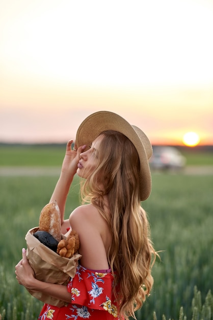 Eine Blondine in rotem Kleid und Strohhut mit französischem Brot in den Händen auf einem Weizenfeld bei Sonnenuntergang