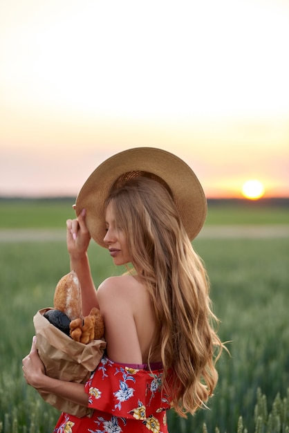 Eine Blondine in rotem Kleid und Strohhut mit französischem Brot in den Händen auf einem Weizenfeld bei Sonnenuntergang