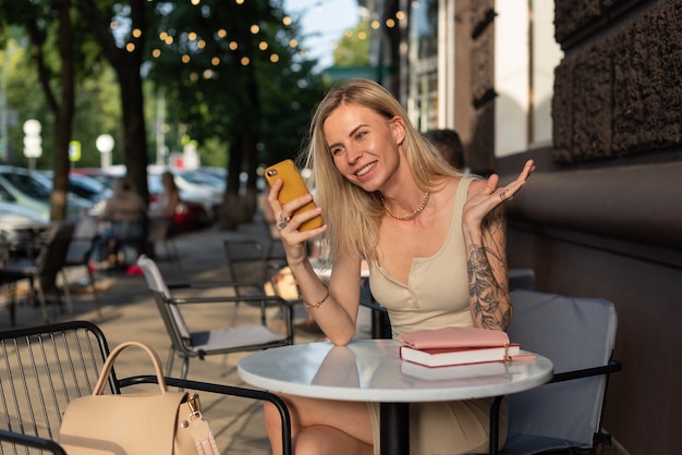 Eine Blondine mit Tattoo auf dem Arm sitzt in einem Sommercafé und telefoniert emotional