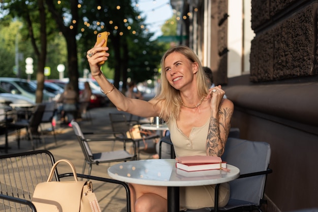 Eine Blondine mit Tattoo auf dem Arm sitzt in einem Sommercafé und telefoniert emotional