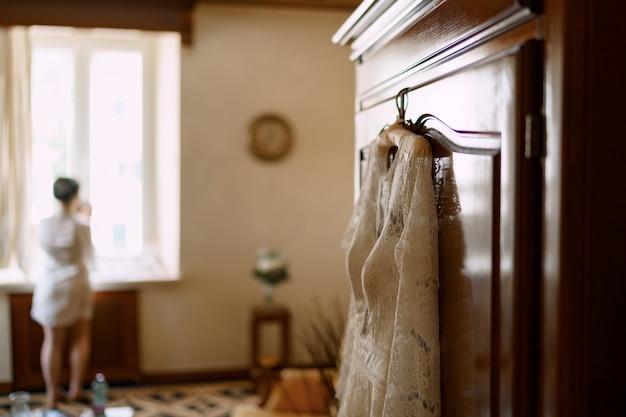 Eine Braut im Gewand steht am Fenster in einem Hotelzimmer ein Hochzeitskleid hängt an der Schranktür