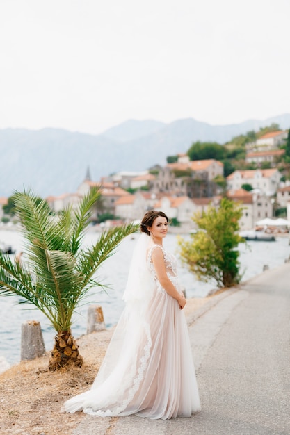 Eine Braut in einem zarten Kleid und einem Schleier steht auf dem Pier in der Nähe der Altstadt von Perast