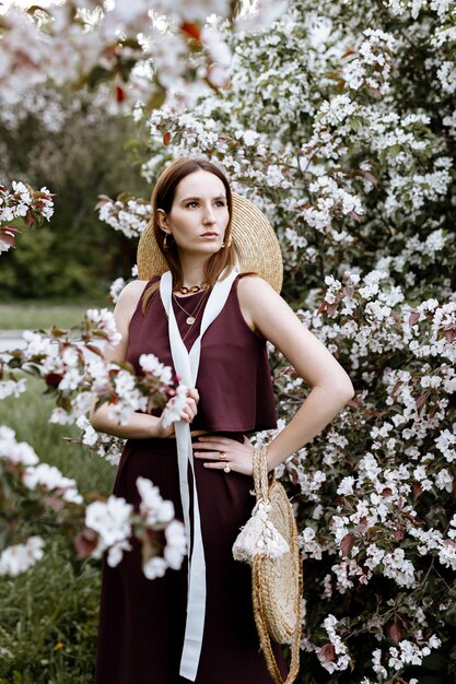 Eine brünette Frau in einem eleganten Anzug steht in der Nähe von Apfelbäumen in voller Blüte an einem warmen Sommertag.