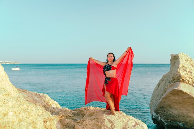 Eine brünette Frau in einem roten orientalischen Tanzkostüm tanzt mit einem Schal auf einem Felsen vor dem Hintergrund des Meeres.