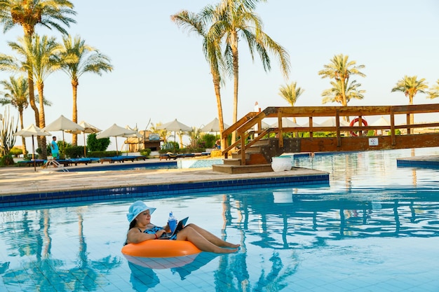 Eine brünette Frau mit Sonnenhut in einem Schwimmbad in einem Schwimmkreis mit einem Cocktail