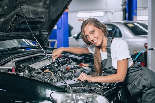 Eine Brünette in einem schwarzen Overall und einem weißen T-Shirt neben der offenen Motorhaube des schwarzen Autos. Junge Frau in der Garage lächelt in die Kamera. Autoreparaturkonzept