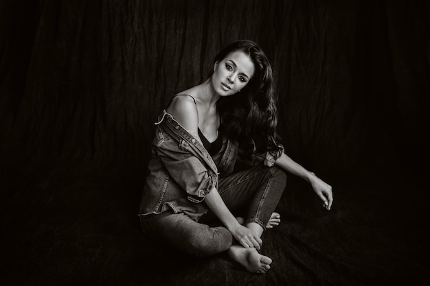 Eine Brünette mit langen Haaren posiert im Studio auf einem schwarzen Hintergrund auf dem Boden sitzend. Schwarz-Weiß-Foto